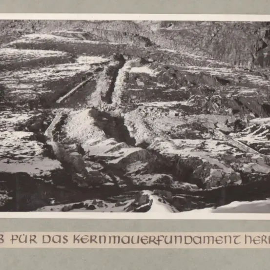 Ein graustufen Foto einer rauen Berglandschaft mit sichtbarer Erosion und Felsformationen. Schnee bedeckt teilweise den Boden.