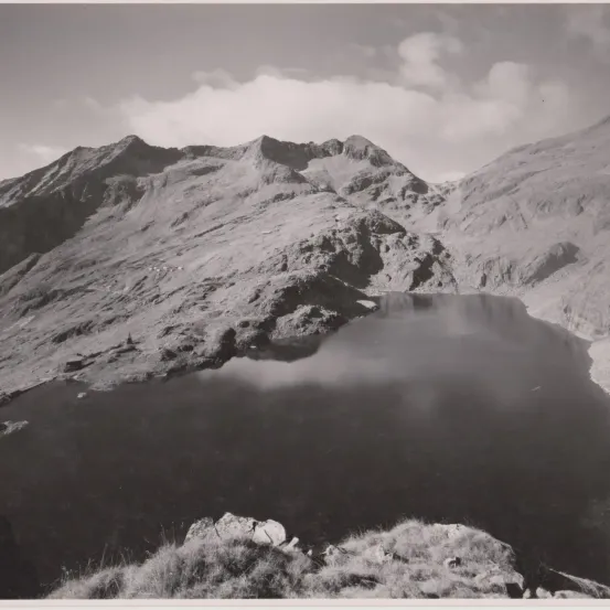 Ein Schwarz-Weiß-Foto einer Gebirgslandschaft mit einem friedlichen See, der zwischen den Gipfeln unter einem bewölkten Himmel liegt.