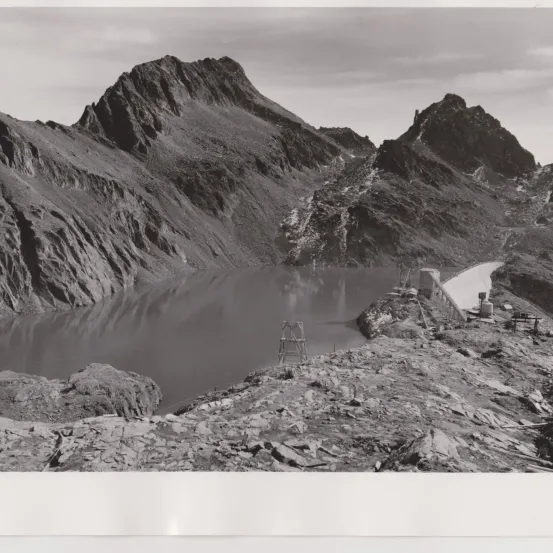 Schwarz-Weiß-Foto eines Bergsees mit felsigen Klippen und einem Damm im Hintergrund.