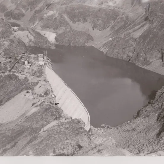 Ein Schwarz-Weiß-Bild eines großen Damms mit einem Fluss, der hindurchfließt. Berge umgeben das Gebiet, und der Bau des Damms ist sichtbar.