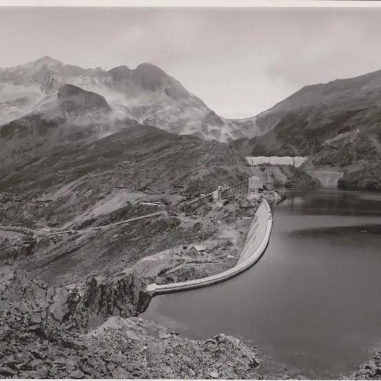 Schwarz-Weiß-Foto einer Berglandschaft mit einem Damm. Im Hintergrund sind schneebedeckte Berge zu sehen.