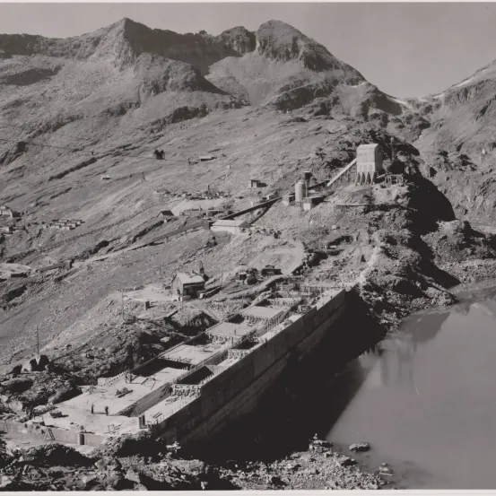 Ein Schwarz-Weiß-Bild zeigt ein bergiges Gebiet mit einem gebauten Damm. Es gibt mehrere Gebäude und Maschinen am Berghang, und ein Gewässer befindet sich vor dem Damm.