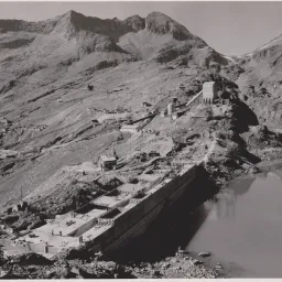 Ein Schwarz-Weiß-Bild zeigt ein bergiges Gebiet mit einem gebauten Damm. Es gibt mehrere Gebäude und Maschinen am Berghang, und ein Gewässer befindet sich vor dem Damm.