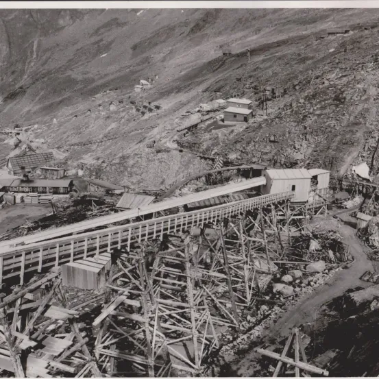 Ein Schwarz-Weiß-Foto zeigt eine Luftaufnahme eines bergigen Bergbaugebiets mit mehreren Gebäuden und einer langen Holzbrücke über Schutt. Im Hintergrund sind Gebäude und eine Bahnstrecke zu sehen.
