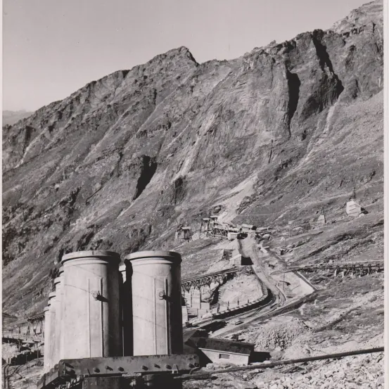 Ein Schwarz-Weiß-Foto zeigt zwei große zylindrische Strukturen im Vordergrund mit einem bergigen Hintergrund und einem gewundenen Pfad auf dem Berg.