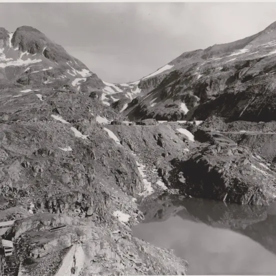 Ein Schwarz-Weiß-Foto zeigt eine Berglandschaft mit einem Fluss, der durch das Tal fließt, flankiert von steilen Klippen und schneebedeckten Gipfeln. Eine Straße windet sich durch die Berge, und es gibt Anzeichen von Bauaktivitäten auf der linken Seite.