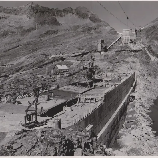 Ein Schwarz-Weiß-Bild zeigt eine Baustelle für einen Damm auf einem Berg mit felsigem Gelände. Gebäude und Maschinen sind verstreut.