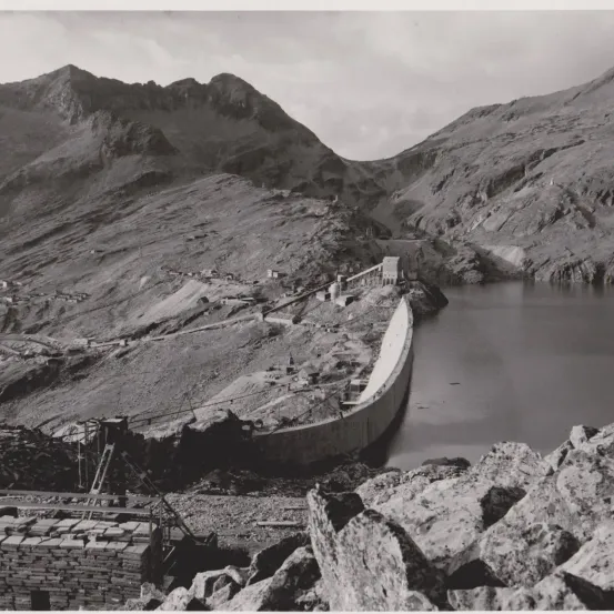 Ein Schwarz-Weiß-Foto zeigt ein bergiges Gebiet mit einem im Bau befindlichen Damm. Der Damm erstreckt sich über einen Fluss, mit Steinhaufen und einer kleinen Struktur auf der linken Seite. Im Hintergrund erheben sich Berge.