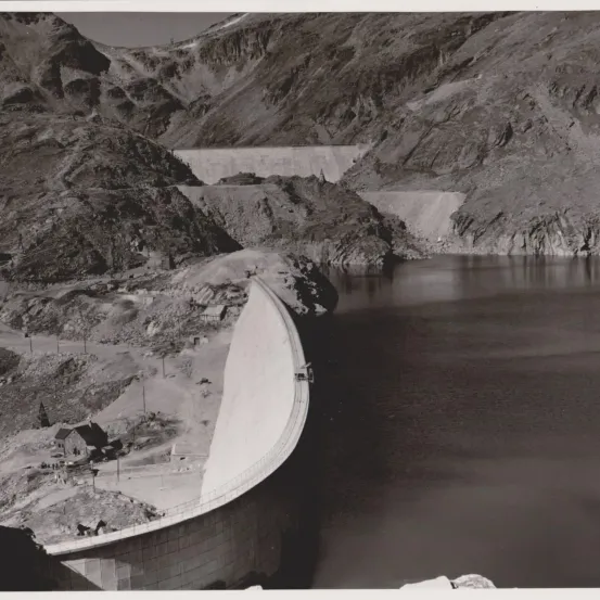 Ein Schwarz-Weiß-Foto zeigt einen im Bau befindlichen Damm in einer Bergregion. Berge umgeben den Damm, und Wasser füllt das Gebiet dahinter.