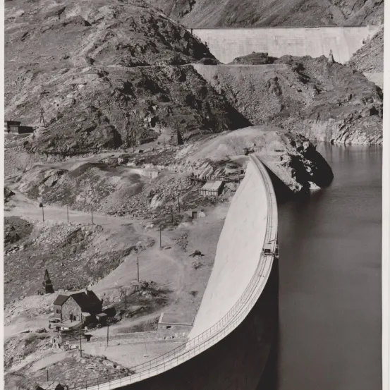 Schwarz-Weiß-Bild eines großen Damms im Bau mit einer Bergkette im Hintergrund.