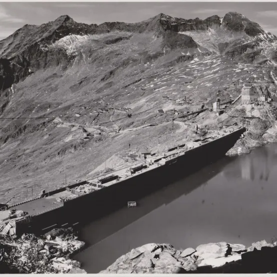 Ein Schwarz-Weiß-Foto zeigt einen Berg mit einem Damm an seiner Basis. Gebäude und eine Straße sind am Berghang sichtbar.
