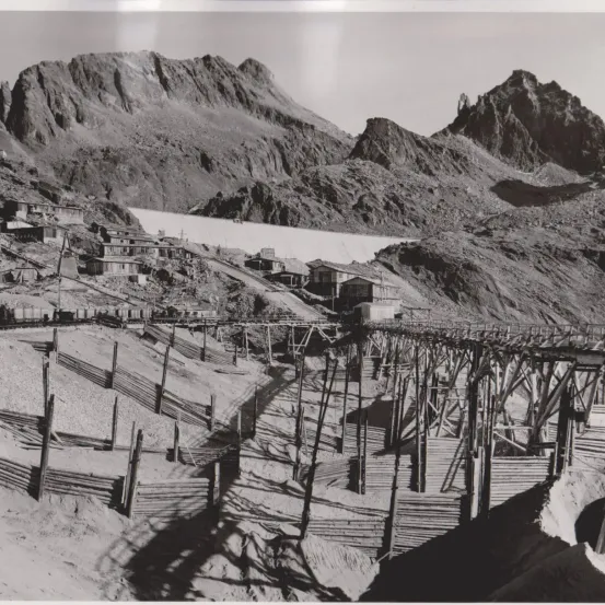 Ein Schwarz-Weiß-Foto zeigt ein bergiges Gebiet mit mehreren kleinen Gebäuden und einer langen Bahnstrecke. Die Berge sind rau mit steilen Hängen. Ein Damm oder Stausee ist hinter den Gebäuden sichtbar.