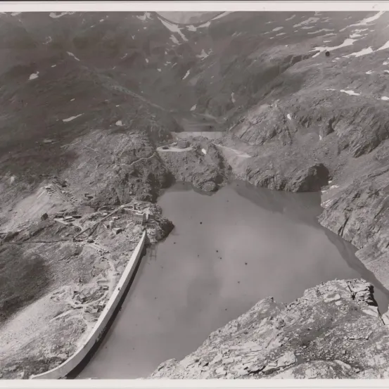 Ein Schwarz-Weiß-Foto einer Berglandschaft mit einem großen Gewässer, möglicherweise einem Stausee, umgeben von rauem Gelände und entfernten schneebedeckten Gipfeln.