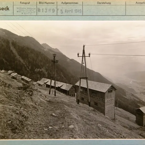 Ein altes Foto zeigt ein bergiges Gebiet mit mehreren kleinen Häusern, Stromleitungen und einer Seilbahn. Das Bild ist vom 5. April 1949 datiert.