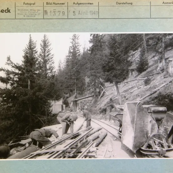 Ein Schwarz-Weiß-Foto zeigt eine Baustelle mit Arbeitern. Ein Arbeiter ist links und zwei sind rechts und arbeiten an einer Eisenbahnstrecke. Holzstämme liegen auf dem Boden. Bäume und ein Gebäude sind auf der linken Seite sichtbar.