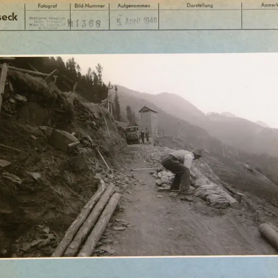 Ein Schwarz-Weiß-Foto zeigt einen Mann, der auf einem Weg mit mehreren Baumstämmen und einem Fahrzeug im Hintergrund arbeitet. Das Bild ist mit dem Namen des Fotografen und dem Datum beschriftet.