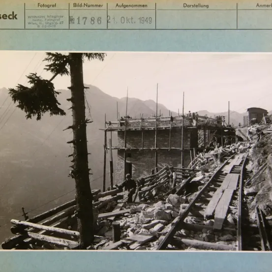 Ein Schwarz-Weiß-Foto zeigt eine Baustelle mit einem im Bau befindlichen Holzgebäude, umgeben von einer Bergkulisse. Das Bild enthält Text auf Deutsch und umfasst eine Fotonummer, Datum und weitere Details.