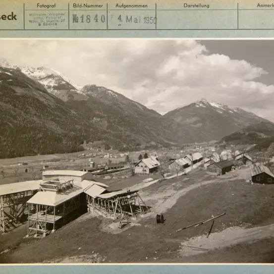 Ein Foto vom 4. Mai 1950 zeigt eine bergige Landschaft mit mehreren Gebäuden, darunter eine Baustelle, und einer kurvenreichen Straße. Das Bild ist mit dem Namen des Fotografen, Wilhelm Wagner, und einer eindeutigen Bildnummer versehen.