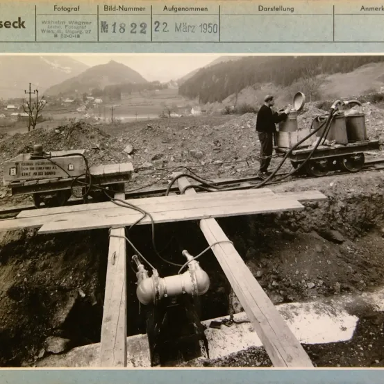 Schwarz-Weiß-Foto zeigt einen Mann, der Wasser in einen großen Behälter auf einem Gleis gießt. Ein kleiner Zug mit Fässern ist im Hintergrund. Ein Ventil und Rohre sind im Boden vergraben. Datum ist 22. März 1950.