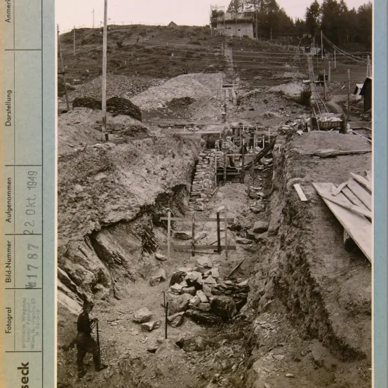 Ein Schwarz-Weiß-Foto zeigt eine Baustelle mit Haufen von Steinen und einem Mann im Vordergrund. Das Jahr 1949 ist auf der linken Seite sichtbar.