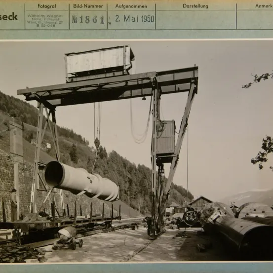 Ein Foto von einer großen Rohrleitung, die von einem Kran gehoben wird. Die Rohrleitung wird von einer Gruppe von Arbeitern bewegt. Das Datum des Fotos ist der 2. Mai 1950.