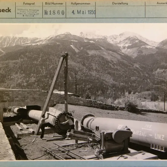 Schwarz-Weiß-Foto, das den Bau einer großen Rohrleitung oder eines Tunnels mit Arbeitern und Maschinen zeigt, datiert auf den 4. Mai 1950, mit einem Gebirgshintergrund.
