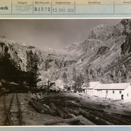 Schwarz-Weiß-Foto einer bergigen Eisenbahnbaustelle mit einem kleinen Gebäude und Holzstapeln. Das Foto hat eine Beschriftung, die lautet 'Wilhelm Wagner Techni. Fotogr. Wien, III. Ungar. 27 B 520-0-1e.' Das Foto wurde am 13. Oktober 1950 aufgenommen.