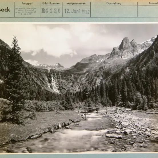 Schwarz-Weiß-Foto einer Berglandschaft. Es zeigt eine kleine Brücke über einen fließenden Fluss, umgeben von Bäumen und felsigem Gelände. Das Bild wurde am 12. Juni 1952 aufgenommen.