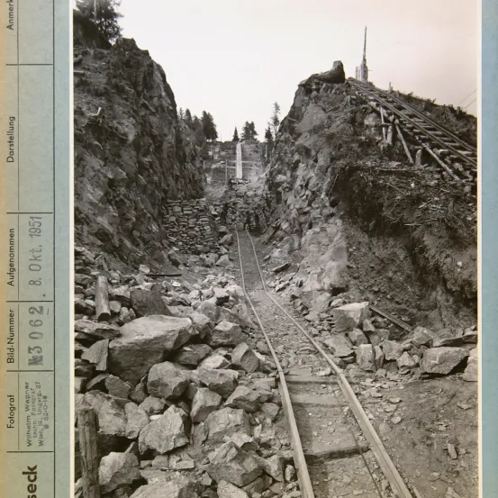 Ein Schwarz-Weiß-Foto zeigt einen felsigen Berg mit einer Eisenbahnstrecke, die hinaufführt. Die Strecke ist von Felsen umgeben, und es gibt einen steilen Anstieg. In der Ferne steht ein Turm auf dem Berggipfel.