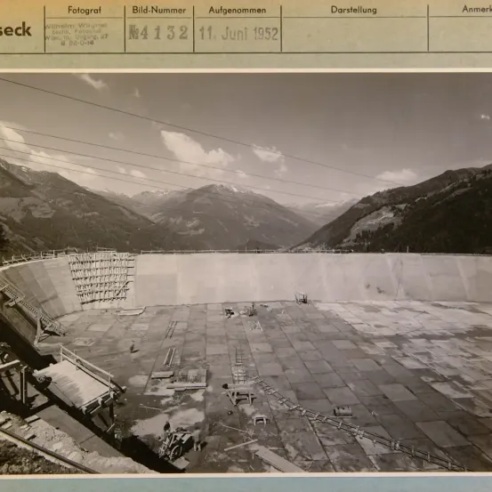Schwarz-Weiß-Foto zeigt eine Baustelle mit Bergen im Hintergrund. Die Baustelle hat einen großen, teilweise gebauten Damm. Das Datum der Aufnahme ist der 11. Juni 1952.