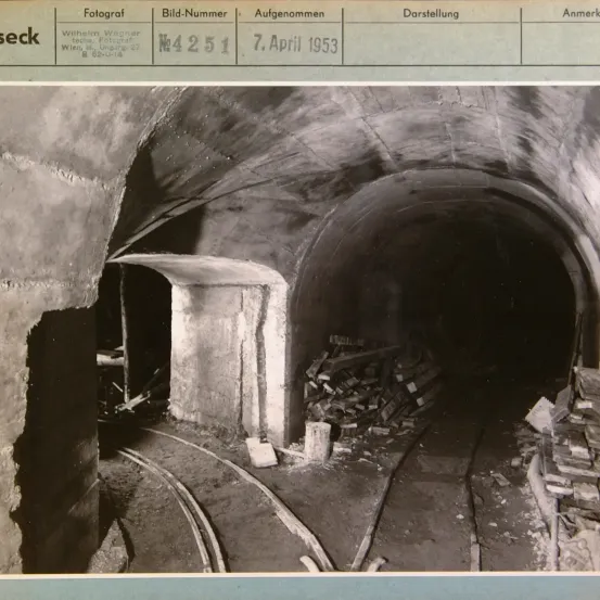 Ein Schwarz-Weiß-Foto zeigt das Innere eines Tunnels mit Holzstapeln und Gleisen auf dem Boden. Es ist vom 7. April 1953 und wurde von Wilhelm Wagner in Wien aufgenommen.