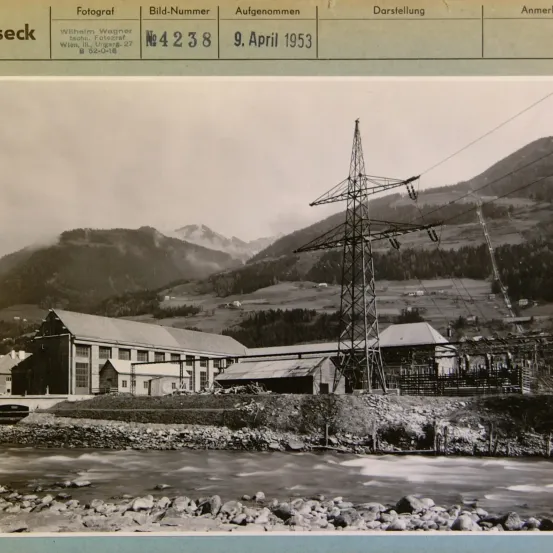 Ein Schwarz-Weiß-Foto einer bergigen Gegend mit einem Fluss und einem Kraftwerk. Das Bild hat eine Beschriftung mit dem Datum '9. April 1953'. Es zeigt ein Gebäude mit vielen Fenstern.