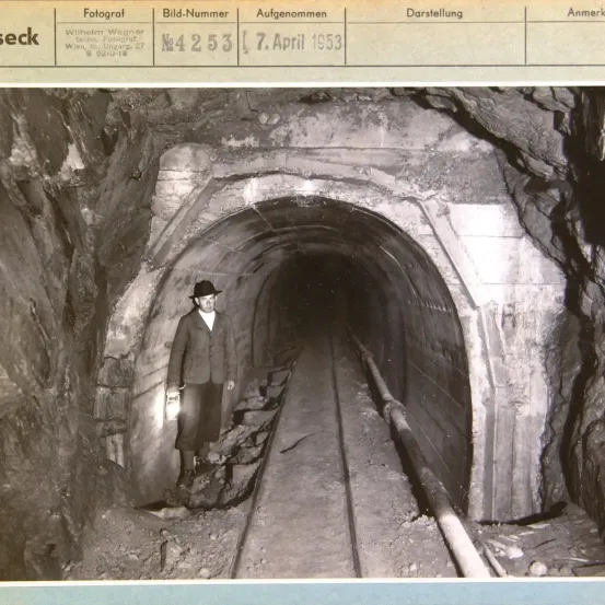 Schwarz-Weiß-Foto eines Mannes, der in einem Tunnel steht. Der Tunnel hat einen gebogenen Pfad und eine Metallrohrleitung auf der rechten Seite. Der Mann trägt einen Hut und hält eine Laterne.