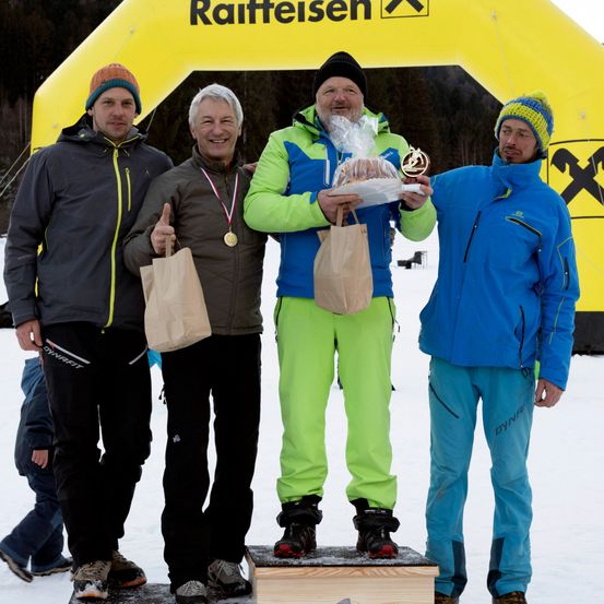 Vier Männer stehen auf einem Podium, einer hält eine Trophäe und einen Kuchen. Sie tragen Winterkleidung. Hinter ihnen ist ein gelber Banner mit dem Wort Raiffeisen.
