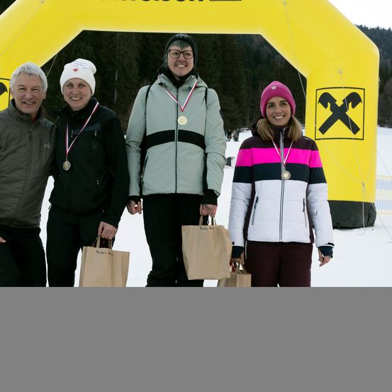 Vier Menschen stehen im Schnee, lächelnd, in Winterkleidung und mit Medaillen. Sie halten braune Papiertüten. Hinter ihnen steht ein gelber Bogen mit einem schwarzen X-Zeichen.