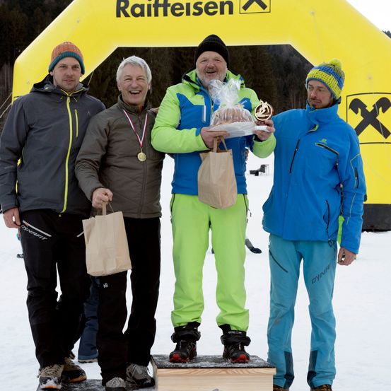 Vier Männer stehen auf einem Podest im Schnee. Ein Mann hält eine Trophäe und einen Kuchen, während andere Papiertüten halten. Sie tragen Winterkleidung und lächeln. Dahinter befindet sich eine gelbe aufblasbare Struktur mit dem Wort 'Raiffeisen'.