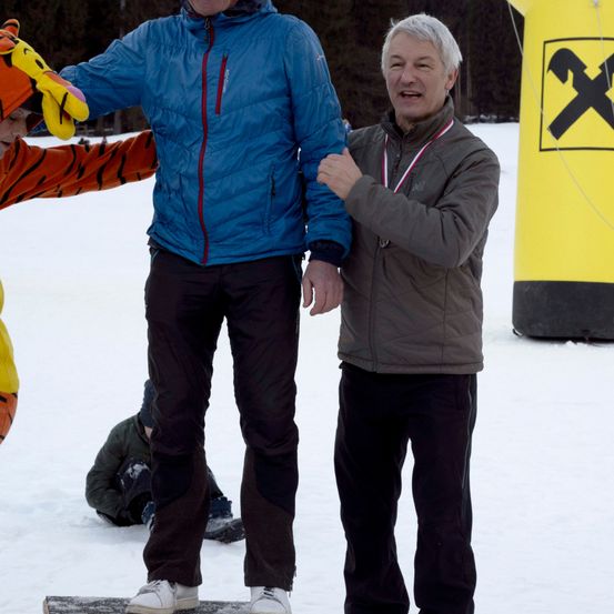 Zwei Männer stehen auf einem verschneiten Feld, einer trägt eine blaue Jacke und der andere eine braune Jacke mit einer Medaille. Eine Person in einem Tigerkostüm steht hinter ihnen. Ein großes gelbes aufblasbares Objekt ist im Hintergrund.