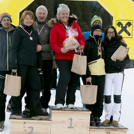 Eine Gruppe von Menschen, möglicherweise Gewinner, steht auf einem verschneiten Podest mit Medaillen und Papiertüten. Das Podest hat die Zahlen 1, 2 und 3. Sie tragen Winterkleidung und halten Papiertüten. Ein gelber Banner mit einem X-Zeichen steht hinter ihnen.