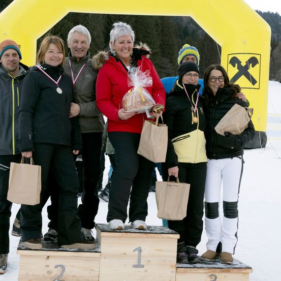 Eine Gruppe von Menschen steht auf einem Podium in einem verschneiten Bereich. Eine Frau in einer roten Jacke hält ein verpacktes Geschenk. Sie tragen alle Medaillen und halten Papiertüten. Dahinter steht ein gelber Bogen mit einem großen X.
