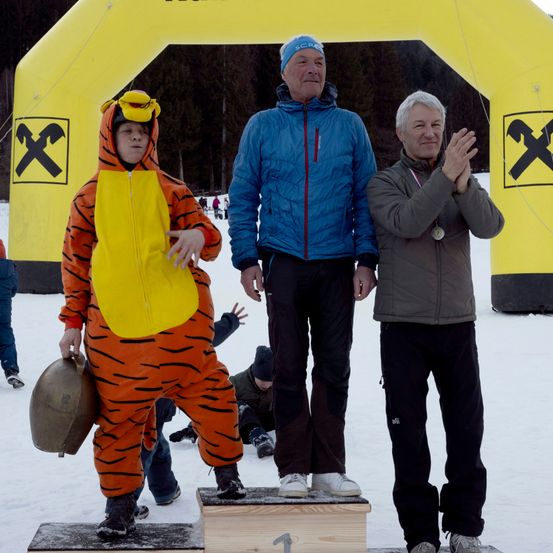 Drei Männer auf einem verschneiten Podest, einer in einem Tigerkostüm, einer in einem blauen Jackett und einer in einem braunen Jackett, lächeln und klatschen. Dahinter ein gelber Bogen mit einem X-Zeichen.