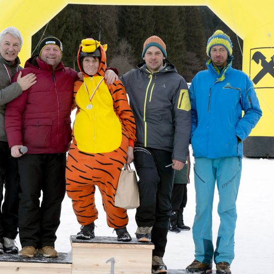 Fünf Personen stehen auf einem verschneiten Podest, eine Person in einem Tigerkostüm mit einer Medaille um den Hals. Dahinter befindet sich eine gelbe Struktur mit einem Logo.