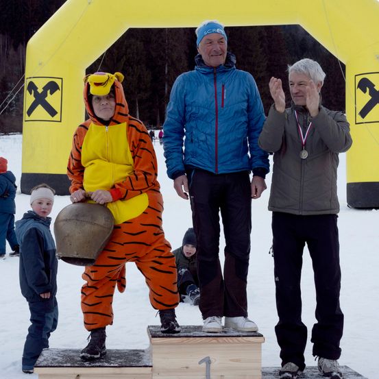 Drei Personen stehen auf einem Podest in einem verschneiten Bereich. Der Mann in der Mitte hält eine Glocke. Der Mann auf der linken Seite trägt ein Tigerkostüm. Der Mann auf der rechten Seite klatscht. Dahinter steht ein gelber Bogen.