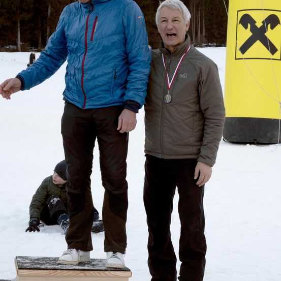 Zwei Männer stehen auf einer verschneiten Oberfläche, einer trägt eine blaue Jacke, der andere eine braune Jacke. Sie haben beide Medaillen um den Hals. Hinter ihnen ist ein gelbes Objekt mit einem schwarzen X zu sehen. Ein Kind sitzt im Hintergrund auf dem Schnee.