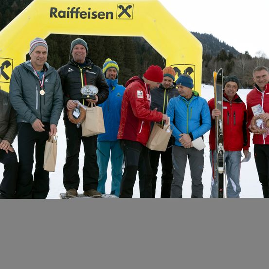 Eine Gruppe von Männern in Winterkleidung steht vor einem gelben Bogen mit Raiffeisen-Logo. Sie halten verschiedene Gegenstände wie Trophäen, Papiertüten und Skier.