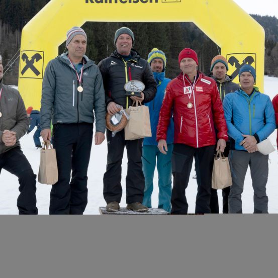 Mehrere Männer stehen in einem verschneiten Bereich, wahrscheinlich nach einem Wettbewerb. Sie tragen Winterjacken, einige halten Papiertüten, und ein Mann hält eine Trophäe. Hinter ihnen steht ein gelber Bogen mit einem Logo.
