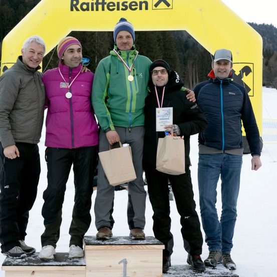Fünf Männer stehen auf einem Podest in Winterkleidung, halten Medaillen und Papiertüten. Hinter ihnen steht ein gelber Bogen mit dem Wort Raiffeisen. Die Männer tragen Jacken, Mützen und Turnschuhe und stehen auf einer verschneiten Oberfläche.