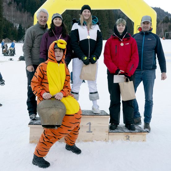 Eine Gruppe von Menschen in Winterkleidung steht auf einem verschneiten Podest mit einem gelben Bogen im Hintergrund. Sie halten Medaillen und Papiertüten.