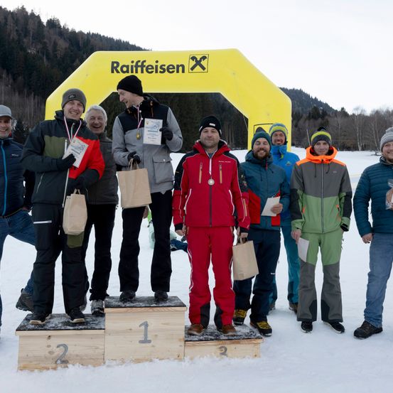 Eine Gruppe von Männern auf einem verschneiten Feld mit einem gelben Bogen, auf dem Raiffeisen steht. Sie halten Papiere und Taschen. Drei Männer stehen auf Podesten mit den Nummern 1, 2 und 3.