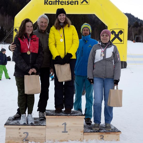 Fünf Personen stehen auf einem verschneiten Podest und tragen Medaillen. Eine Frau in einer gelben Jacke steht auf dem Siegerpodest. Ein Mann und eine Frau stehen auf dem zweiten und dritten Podest. Hinter ihnen ist ein gelbes Banner mit dem Wort Raiffeisen.
