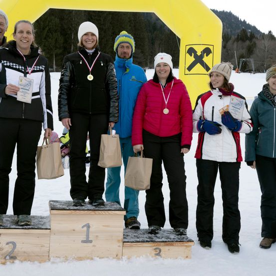 Fünf Personen stehen auf einem verschneiten Feld und halten Medaillen und Papiertüten. Sie tragen Winterkleidung. Drei von ihnen stehen auf einem Podest mit den Nummern 1, 2 und 3. Hinter ihnen befindet sich ein gelber Bogen mit einem X-Symbol.